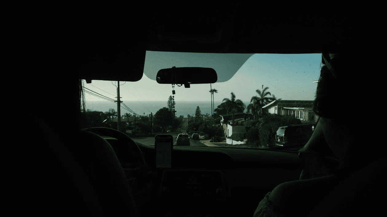 Inside view of a car with two passengers, a phone mounted on the dashboard, and a coastal road ahead, representing a ride-sharing scenario.